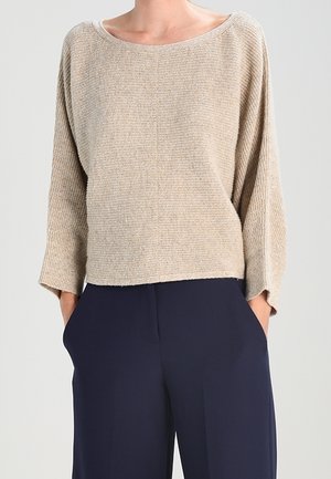 Beige textured long-sleeve sweater with wide neckline paired with loose-fitting navy blue pants, hands in pockets.