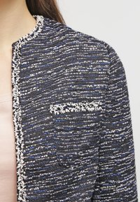 Blå och svart strukturerad jacka med ett vävt mönster, med en vit kant längs halsringningen och en liten ficka på bröstet.