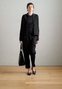 Cardigan noir avec boutons, haut noir et pantalon sombre. Un sac à main noir et des chaussures à pois complètent la tenue. Design simple et texturé.