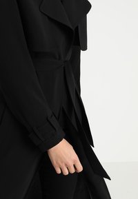 Zbliżenie na osobę w czarnym płaszczu typu trench z paskiem i detalami klamry na rękawie, z widoczną lewą ręką.