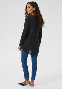 Black long-sleeve shirt with a textured finish, featuring a curved hem and side slits, paired with blue skinny jeans and black heeled shoes.