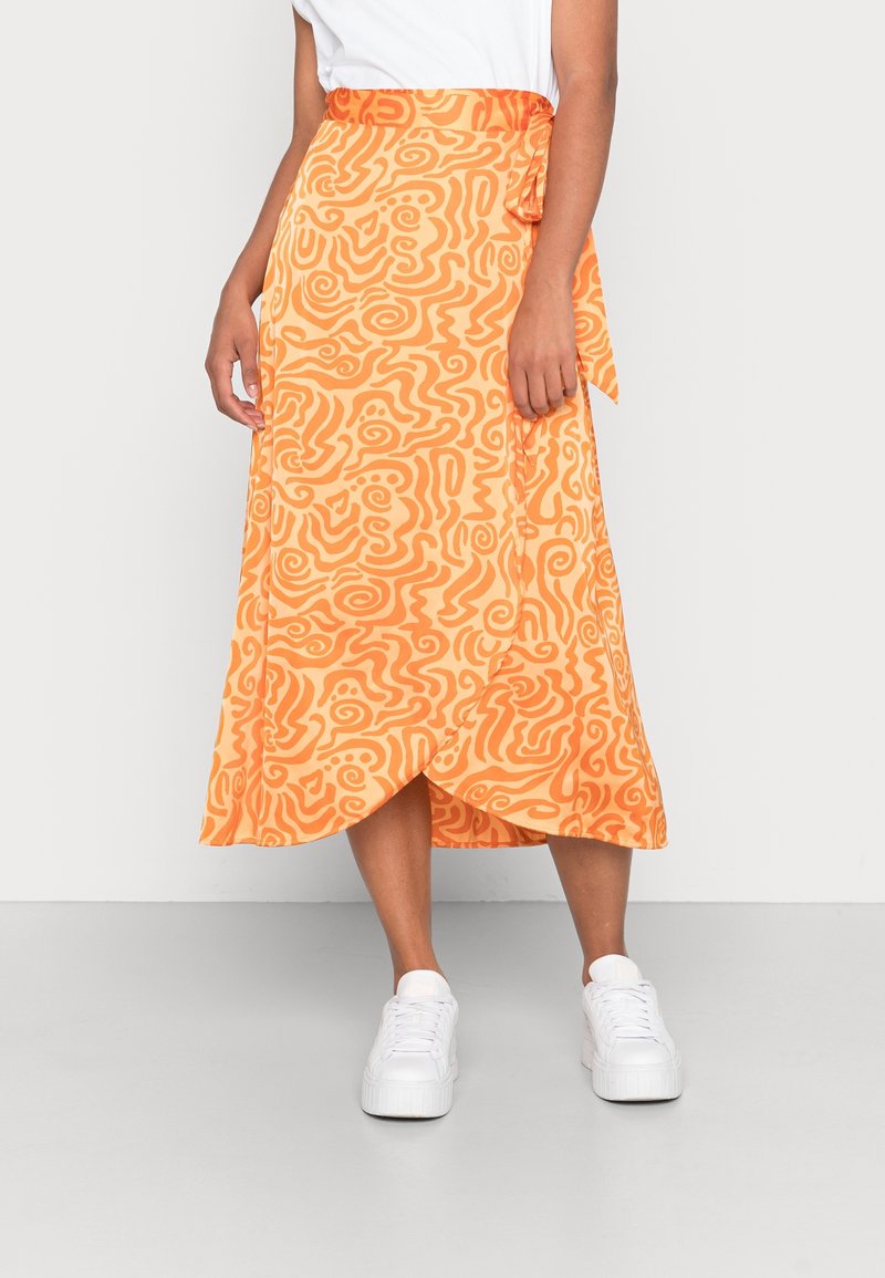 Femme portant une jupe midi croisée avec des motifs abstraits orange et des baskets blanches, se tenant devant un fond clair.