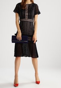 Kvinna i svart spets-midi klänning som håller en marinblå clutch, bär röda högklackade skor, stående mot en enkel vit bakgrund.