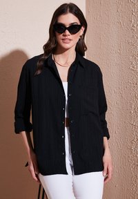 Black ribbed button-up shirt with a chest pocket, long sleeves rolled to the elbows, paired with white pants. Accents include sunglasses and jewelry.