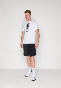 Fehér, rövidujjú Nike póló fekete NBA logóval, sötétszürke sorttal és fehér sportcipővel párosítva. Egyszerű, sportos dizájn.