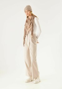 Bonnet en tricot épais beige et châle avec franges, associés à un pantalon large crème et des bottes à plateforme blanches. Design et textures minimalistes.
