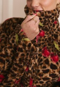 Kurtka z futra sztucznego w leopardzie wzór z zielonymi i czerwonymi akcentami kwiatowymi, wysoki kołnierz, miękka tekstura oraz zbliżenie na rękę trzymającą kołnierz.