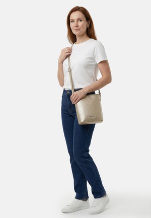 Femme portant un t-shirt blanc, un jean bleu, des baskets blanches, tenant un sac bandoulière doré avec une sangle ajustable sur un fond uni.