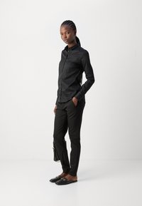 Camicia nera con bottoni in tessuto morbido, abbinata a pantaloni neri su misura e scarpe slip-on nere con dettagli in hardware dorato.