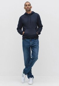 Marinblå tröja med huva och framficka, med snörning, i kombination med ljusblå jeans och vita sneakers med gröna accenter.