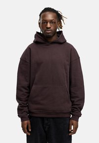 Brauner Kapuzenpullover mit einer Fronttasche, lockerer Passform, gerippte Bündchen und weicher, strukturierter Stoff. Getragen mit schwarzen Jeanshosen.