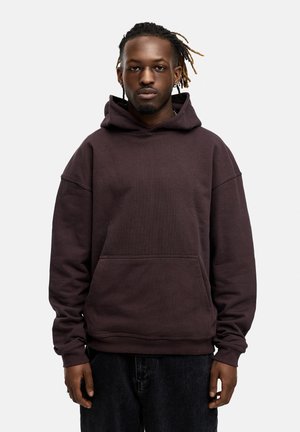Brown hooded sweatshirt with a front pocket, loose fit, ribbed cuffs, and soft, textured fabric. Worn with black denim pants.