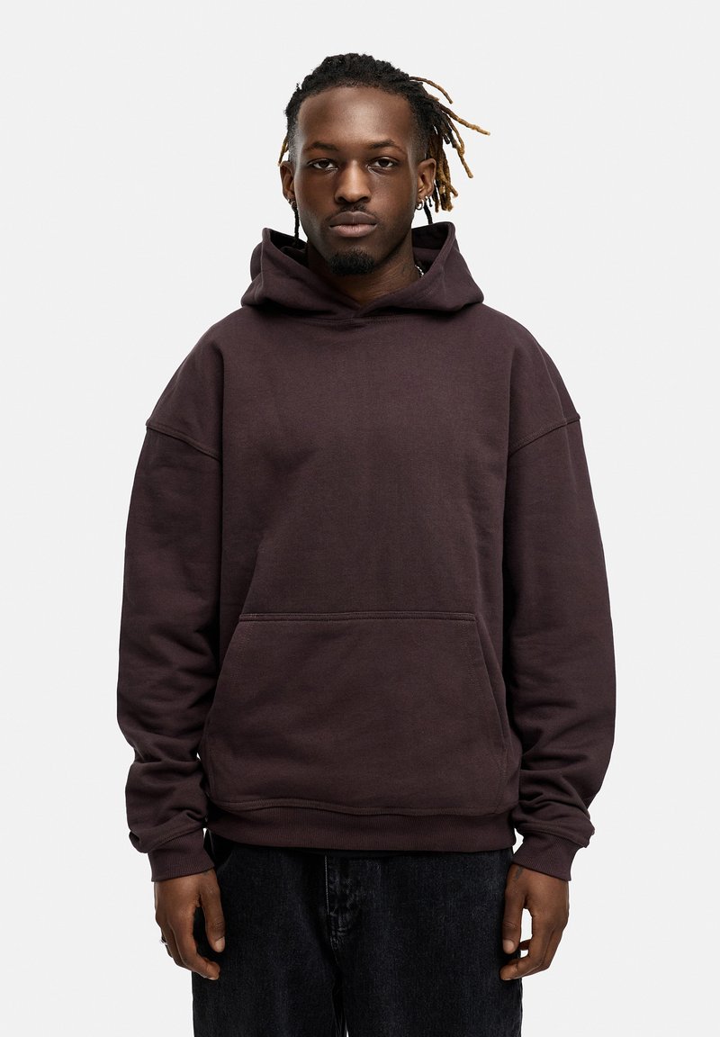 Brauner Kapuzenpullover mit einer Fronttasche, lockerer Passform, gerippte Bündchen und weicher, strukturierter Stoff. Getragen mit schwarzen Jeanshosen.