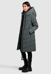 Vrouw in een lange donkergrijze gewatteerde winterjas met capuchon, zwarte leggings en zwarte chunky laarzen, profielstaand op een effen achtergrond.