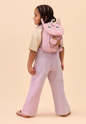 Kind mit geflochtenem Haar, trägt hellrosa Latzhose und einen pinkfarbenen Einhorn-Rucksack, steht seitlich gewandt vor einem beigefarbenen Hintergrund.