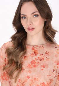 Väldigt rörlig persikofärgad klänning med blommönster i olika nyanser av orange, med rundad halsringning och textil med struktur. Långt, vågigt hår.
