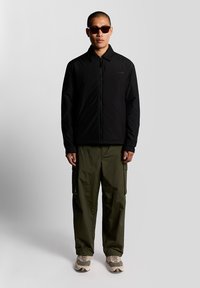 Jachetă neagră cu fermoar, asortată cu pantaloni cargo verde măsliniu. Accesoriile includ ochelari de soare închiși la culoare și adidași bej cu talpă texturată.