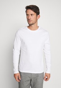 Tricou alb cu mânecă lungă, dintr-un material moale, cu guler rotund și o textură simplă și netedă, purtat cu pantaloni gri cu model.