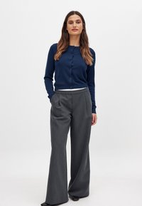 Marineblauer Strick-Cardigan mit Knopfverschluss, kombiniert mit grauen weit geschnittenen Hosen, die ein Nadelstreifenmuster und eine leicht strukturierte Oberfläche aufweisen.