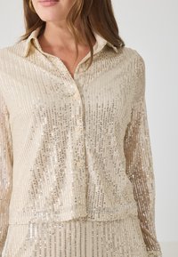 Cămașă bej cu paiete, cu nasturi, cu dungi verticale, textură moale, detaliu la guler și un luciu subtil. Pantalonii coordonați sunt parțial vizibili.