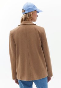 Hnědý blazer z vlněné směsi s přiléhavým střihem, s klopami a jedním zadním roz ventilem, doplněný světle modrou kšiltovkou.