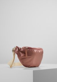 Compact rosegouden satijnen heuptas met rits en beige verstelbare schouderband, geplaatst op een witte voetstuk tegen een grijze achtergrond.