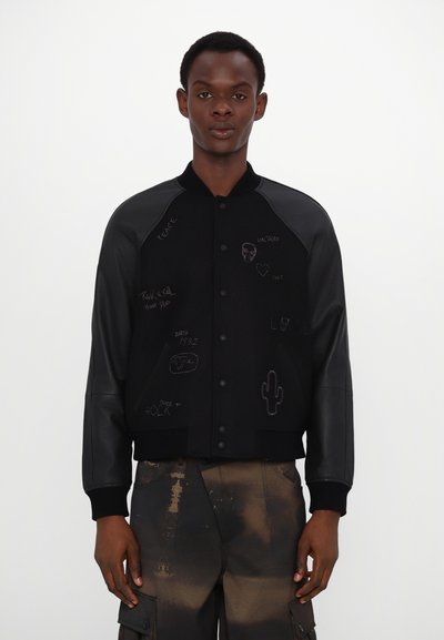 Veste bomber noire avec des manches en cuir, présentant divers designs graphiques et textes à l'avant. Col classique et fermeture à boutons-pression.