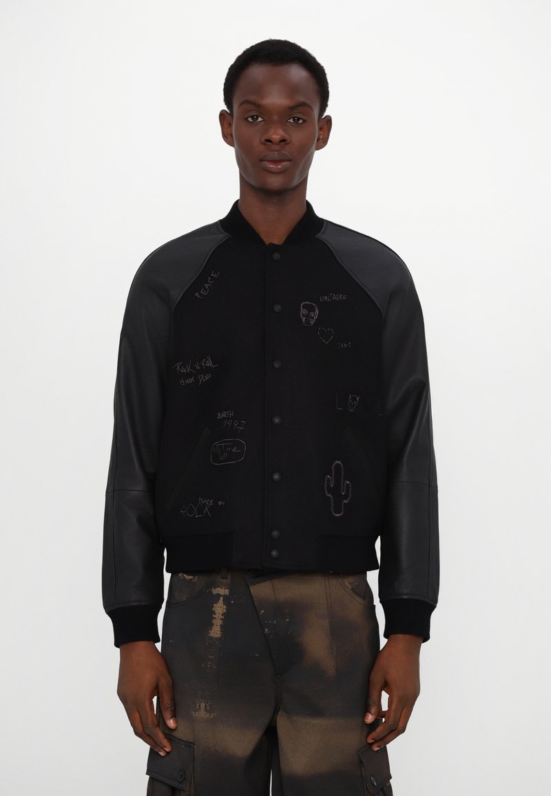 Veste bomber noire avec des manches en cuir, présentant divers designs graphiques et textes à l'avant. Col classique et fermeture à boutons-pression.