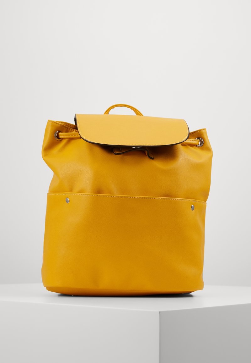 Mochila vegana amarilla de cuero sintético con tapa de solapa, cierre con cordón y bolsillo frontal. Textura suave, forma redondeada y detalles en metal plateado.