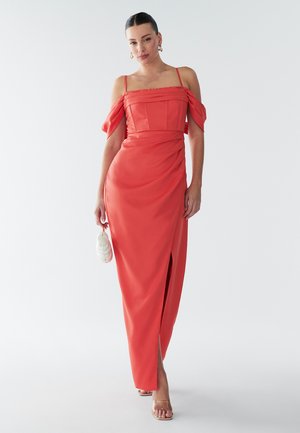 Coral off-shoulder maksimekko, jossa on röyhelökaulus, istuva torso ja sivuaukko, valmistettu sileästä kankaasta. Malli pitää valkoista laukkua.