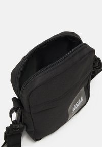 Sac bandoulière en tissu noir avec fermeture éclair, sangle réglable, poche avant et détail du logo. Surface texurée, forme rectangulaire compacte.