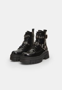 Botas de tobillo decharol negro con una plataforma gruesa, presentan un diseño con cordones, ojales de metal y dos correas ajustables con hebillas.