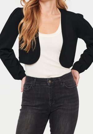 Cárdigan negro corto con mangas abullonadas, usado sobre una camiseta blanca sin mangas, combinado con jeans de mezclilla oscura. Presenta una textura de punto suave.