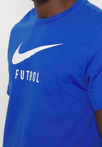 Modré bavlněné tričko s velkým bílým logem Nike swoosh a slovem "FUTBOL" vytištěným níže tučnými bílými písmeny. Krátké rukávy, kulatý výstřih.