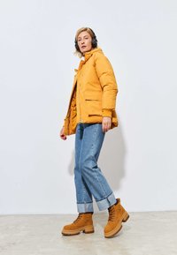 Geel pufferjack met een capuchon, twee voorzakken, gecombineerd met lichtblauwe rechte jeans en tan kleurige veterschoenen met dikke zolen.