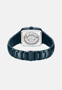 Quadratische, dunkelblaue Edelstahl-Uhr mit transparentem Gehäuseboden, der komplexe Mechanik zeigt. Das Armband hat eine robuste, glatte Oberfläche.