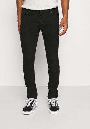 Homme portant un jean noir slim et des baskets noires et blanches à lacets, debout devant un fond clair uni.
