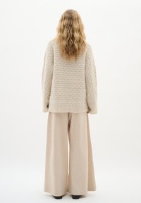 Beige strukturierter Strickpullover mit lockerer Passform und gerippten Bündchen, kombiniert mit hellbeigen Weitbein-Hosen. Model von hinten betrachtet.