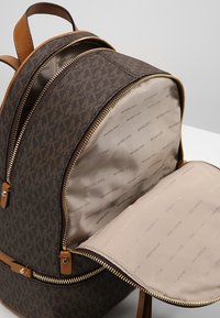 Sac à dos marron à motifs avec des accents en cuir beige, des fermetures à glissière dorées et un intérieur beige doublé du logo de la marque.