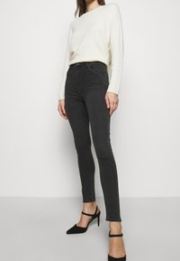 Jeans negros de cintura alta y corte ajustado con una textura suave, combinados con un suéter de punto color crema y zapatos de tacón negros.