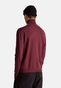 Pullover bordeaux con colletto alto, realizzato in tessuto morbido. Presenta una texture a coste e un design aderente, abbinato a un berretto nero.