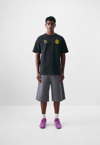 Svart bomull t-skjorte med gult logo, grå denimshorts og rosa sportssko. Modellen står mot en ensfarget bakgrunn.