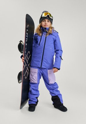 Giacca e pantaloni da snowboard viola con accenti neri e viola chiaro, in posa con una tavola da snowboard, indossando un berretto nero e occhiali da sci.
