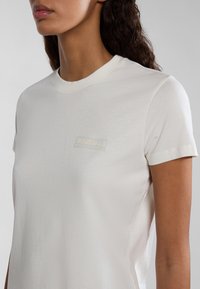 Weißes Baumwoll-T-Shirt mit kurzen Ärmeln, das ein dezentes Logo-Patch auf der Brust aufweist. Glatte Textur und klassisches Rundhalsdesign.