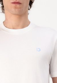Camiseta blanca de algodón con cuello redondo, que presenta un pequeño logo azul en el área del pecho izquierdo. Textura suave y mangas cortas.