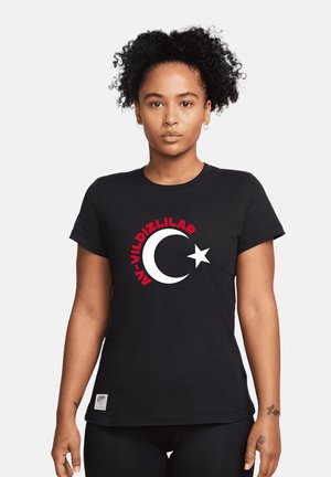 Femme aux cheveux bouclés portant un t-shirt noir avec un croissant blanc et une étoile, avec un texte rouge au-dessus.