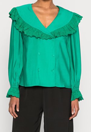 Groene blouse met lange mouwen, grote kanten kraag, dubbele knopen en kanten manchetten, gedragen met een zwarte broek.