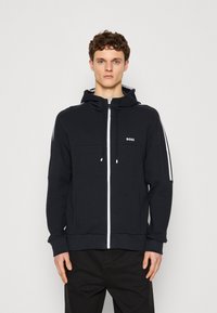 Mörkblå zip-up hoodie med en huva med dragsko, vita sidostråk och en liten logotypdetalj på bröstet. Mjuk, avslappnad tygtextur.