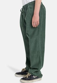 Groene corduroy broek met een elastische tailleband en koord, met verticale ribbels en zijnaad; gedragen met zwart gemusterde sneakers.