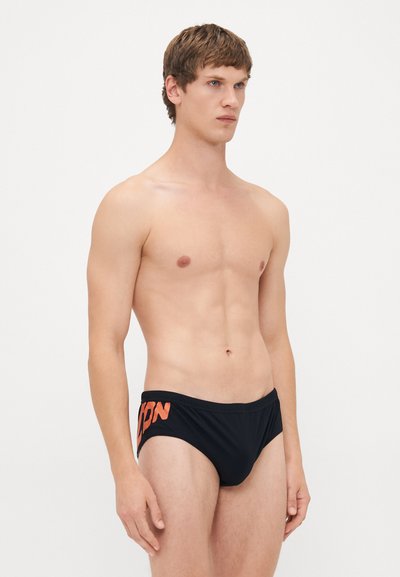 Oben ohne Mann mit kurzen braunen Haaren, der schwarze Badeslips mit orangefarbenem Aufdruck an der Seite trägt, vor einem schlichten weißen Hintergrund stehend.